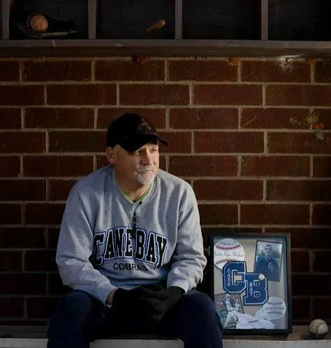 David Shelton sitting in a baseball dugout beside a framed tribute to his son Justin